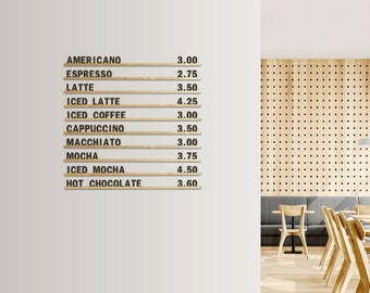 Op de afbeelding: Een witte muur met een houten menu waarop koffiedrankjes en prijzen staan vermeld. Het menu is gemaakt van houten latten met zwarte letters. De muur bevindt zich in een café met houten tafels en stoelen.
