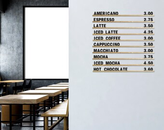 Op de afbeelding: Een witte muur met een houten menu waarop koffiedrankjes en prijzen staan vermeld. Het menu is in zwarte letters geschreven. De muur bevindt zich in een café met houten tafels en stoelen.