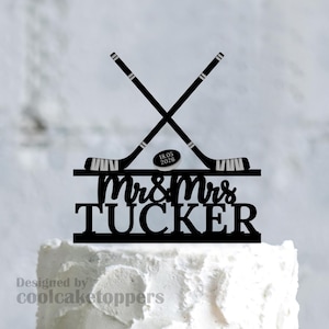 Pode incluir: Topper de bolo preto com tacos de hóquei cruzados, um disco com a data 18.05.2026 e os nomes "Mrs & Mrs Tucker". Desenhado para um bolo de casamento, adiciona um toque desportivo à celebração.