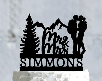 May include: Black silhouette cake topper featuring a couple kissing in front of a mountain range and a pine tree. The text "Mr & Mrs SIMMONS" is below the couple.