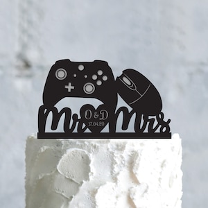 May include: Black and white cake topper featuring a video game controller and a computer mouse with the words "Mr. & Mrs." and the date "O&D 17.04.20".