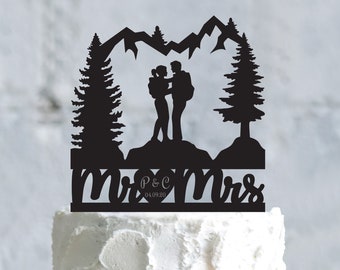 May include: Black silhouette cake topper featuring a couple standing on a mountaintop with trees and mountains in the background. The text "Mr & Mrs" with a heart and the date "04.09.20" is below the couple.