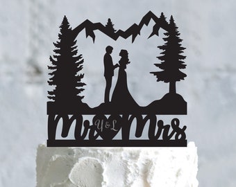 May include: Black silhouette cake topper featuring a couple standing in front of a mountain range with two pine trees. The text "Mr & Mrs" is written below the couple.