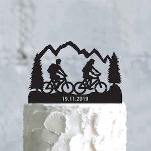 May include: Black cake topper featuring silhouettes of two cyclists riding mountain bikes, with a mountain range and pine trees in the background. The date "19.11.2019" is displayed below the cyclists. The topper is placed on a white cake.
