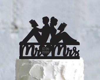 Op de afbeelding: Zwarte silhouet-taarttopper van een stel dat zit en boeken leest. De topper zegt "Mr. & Mrs." met een hart en de datum "17.06.2020" in het midden.