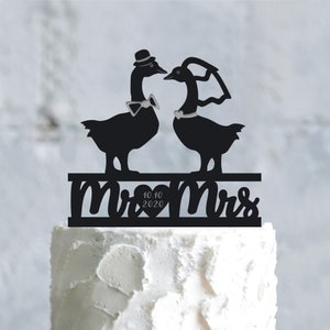 May include: Black silhouette of a bride and groom goose cake topper with the text "Mr & Mrs" and a heart with the date "10.10.2020" on a white cake.