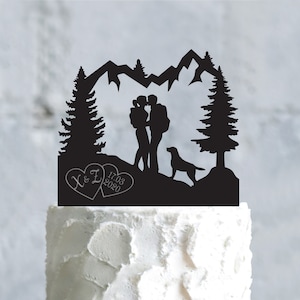 Peut inclure: Topper de gâteau en silhouette noire représentant un couple s'embrassant devant une chaîne de montagnes avec deux sapins et un chien. Le couple se tient la main et l'homme porte un sac à dos. La date 17.03.2020 et les initiales X & L sont inscrites dans deux cœurs.