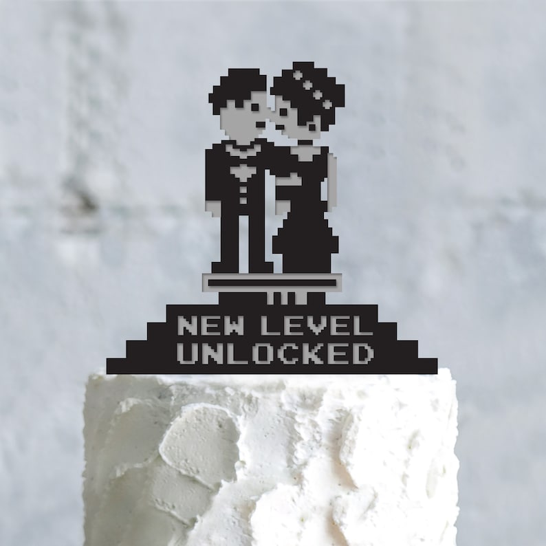 May include: Black pixelated wedding cake topper featuring a bride and groom embracing. The text "NEW LEVEL UNLOCKED" is displayed below the couple. The topper is placed on a white frosted cake.