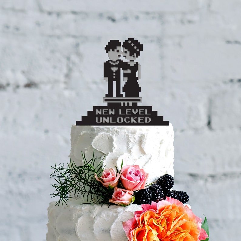 May include: A white tiered wedding cake decorated with pink roses, blackberries, and an orange flower. The cake is topped with a black pixelated cake topper featuring a couple and the text "NEW LEVEL UNLOCKED".