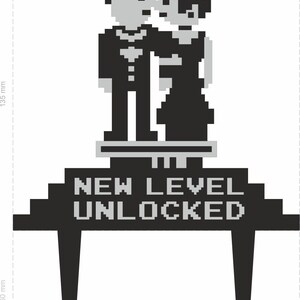 May include: Black and gray pixelated wedding cake topper featuring a couple embracing. The text "NEW LEVEL UNLOCKED" is displayed below the figures. The topper is approximately 5.6 inches wide and 5.3 inches tall.