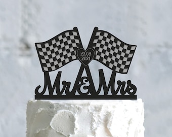 Könnte beinhalten: Schwarzer Tortenaufsatz mit zwei karierten Flaggen, einem Herz mit dem Datum 23.08.2021 und den Worten "Mr & Mrs". Der Aufsatz befindet sich auf einem weiß glasierten Kuchen.