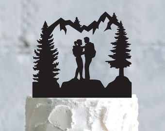 May include: Black silhouette cake topper featuring a couple standing on a rock with a mountain and forest backdrop. The design includes two tall pine trees and a mountain range, perfect for a wedding or anniversary cake.