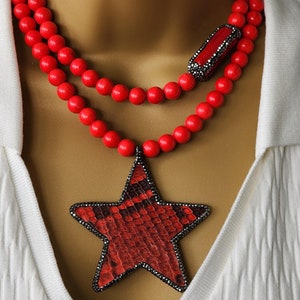 May include: A red beaded necklace with a star pendant. The star pendant is red with a black border and a snakeskin pattern. The necklace is layered and worn over a white V-neck shirt.