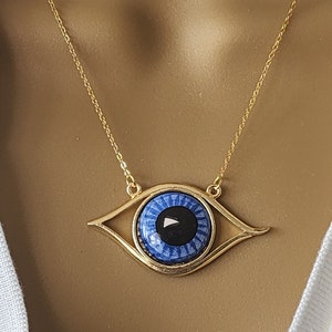 May include: A gold chain necklace with a blue evil eye pendant. The pendant is a large, round eye with a black pupil and white sclera. The eye is surrounded by a gold frame.