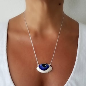 May include: A silver chain necklace with a large blue and white evil eye pendant. The pendant is shaped like an eye with a white iris and a blue pupil.