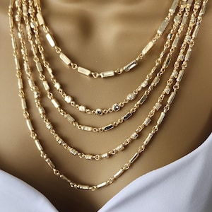 May include: A multi-strand gold-tone necklace. The necklace features several strands of varying designs, including rectangular and beaded elements. The jewelry is displayed against a neutral background, highlighting the warm gold color and intricate details.