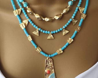 May include: Three-strand necklace featuring turquoise beads and gold-colored metal accents. The top strand has large, irregular beads, the middle strand has small triangle pendants, and the bottom strand has a rectangular pendant with a red and blue design.