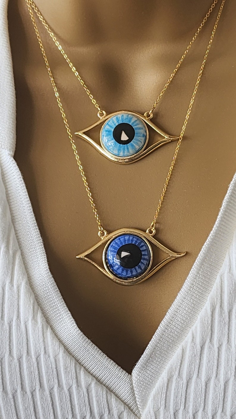 May include: Two gold necklaces with blue evil eye pendants. The top pendant has a light blue eye with a white swirl and a black center. The bottom pendant has a dark blue eye with a white swirl and a black center.