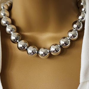 May include: A silver-colored necklace featuring large, round beads. The beads have a reflective, polished surface, creating a shiny appearance. The necklace is displayed against a neutral background, highlighting the jewelry's design.