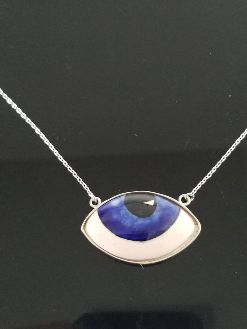 May include: A silver chain necklace with a blue and white evil eye pendant. The pendant is shaped like an eye with a white iris and a blue pupil.
