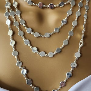 May include: A multi-strand silver-toned necklace featuring numerous small, circular, hammered-texture metal discs linked together by thin silver-toned chains. The necklace drapes in several layers, creating a cascading effect of reflective metallic circles.