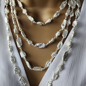 May include: A layered silver necklace with a textured, organic design. The necklace features multiple strands of varying lengths, each adorned with irregularly shaped, metallic beads. The jewelry is displayed against a white background.