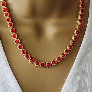 May include: A gold-toned necklace featuring a continuous row of round, vibrant red cabochon stones set in textured metal bezels. The necklace follows a circular path around the neck, showcasing a uniform design of alternating red and gold elements.