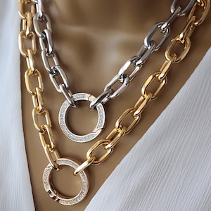 May include: A close-up of two layered necklaces. One necklace is gold-toned, and the other is silver-toned. Both feature large, chunky chain links. Each necklace has a large, round pendant with embedded crystals.