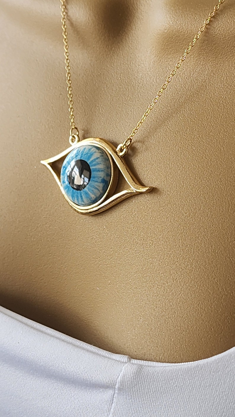May include: Gold chain necklace with a blue eye pendant. The eye is surrounded by a gold frame and has a white and blue swirl pattern.