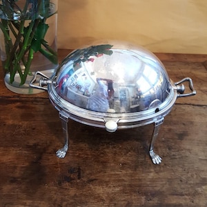 Könnte beinhalten: Eine silberne Servierplatte mit einem gewölbten Deckel und drei Beinen. Die Platte steht auf einem Holztisch mit einem Strauß rosa Rosen in einer Glasvase im Hintergrund.