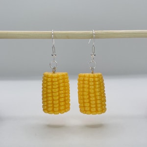May include: A pair of novelty earrings shaped like miniature corn on the cob. The earrings are bright yellow with detailed kernels and hang from silver-colored fish hook ear wires. The earrings are displayed on a wooden dowel.