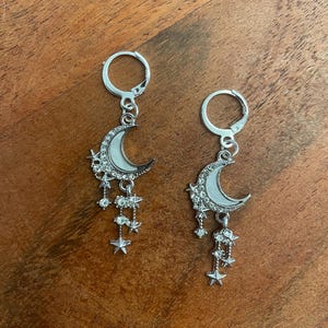 May include: A pair of silver earrings with a crescent moon design, adorned with dangling stars and crystals. The earrings have a lever-back closure. The earrings are displayed on a wooden surface.