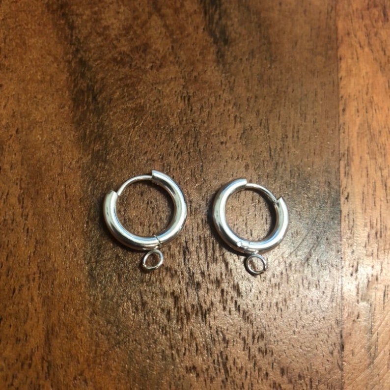 Peut inclure: Une paire de boucles d'oreilles cr&eacute;oles argent&eacute;es, chacune avec une petite boucle en bas pour attacher des breloques. Les boucles d'oreilles sont rondes et brillantes, avec un design simple. Elles sont pos&eacute;es sur une surface en bois.