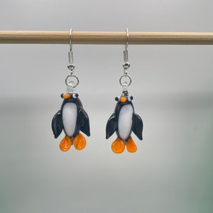 May include: A pair of silver dangle earrings featuring black and white glass penguin charms with orange feet.
