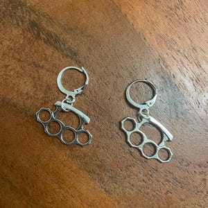 May include: A pair of silver-colored earrings. Each earring features a knuckle duster design with four circular holes. The earrings are suspended from a small, circular hoop. The background is a wooden surface.