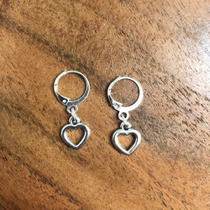 May include: A pair of silver hoop earrings with a dangling heart charm. The heart charm has a cutout design.
