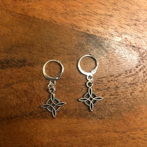Op de afbeelding: Een paar zilverkleurige oorbellen met een Keltisch knoopontwerp. Elke oorbel heeft een klapsluiting en een decoratieve bedel met een ingewikkelde, vierpuntige knoop. De oorbellen worden gepresenteerd op een houten oppervlak.