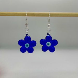 May include: A pair of blue flower-shaped earrings with a central eye design. The earrings are suspended from silver-colored hooks. The flowers have five petals and a small, white and blue eye in the center.