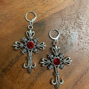 May include: A pair of silver cross earrings with red crystal accents. The earrings are dangling and have a detailed design.