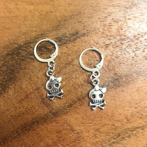 May include: A pair of silver hoop earrings with a skull and crossbones charm with a small bow on top.