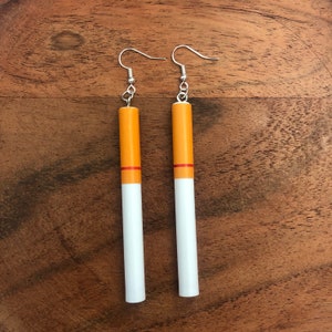 Op de afbeelding: Een paar hangende oorbellen in de vorm van sigaretten. De oorbellen zijn gemaakt van hout en geschilderd om eruit te zien als sigaretten. Het bovenste deel van de oorbellen is oranje, het midden is rood en de onderkant is wit.