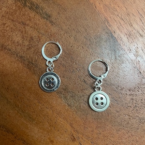 Peut inclure: Une paire de boucles d'oreilles boutons argent&eacute;es. Chaque boucle d'oreille est orn&eacute;e d'un petit bouton rond &agrave; quatre trous, suspendu &agrave; un fermoir &agrave; levier. Les boucles d'oreilles sont pr&eacute;sent&eacute;es sur une surface en bois.