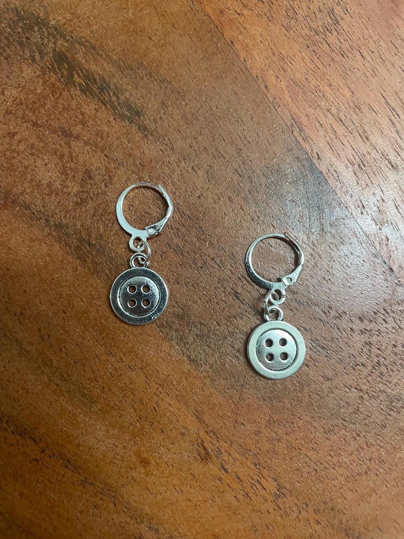 Peut inclure: Une paire de boucles d'oreilles boutons argent&eacute;es. Chaque boucle d'oreille est orn&eacute;e d'un petit bouton rond &agrave; quatre trous, suspendu &agrave; un fermoir &agrave; levier. Les boucles d'oreilles sont pr&eacute;sent&eacute;es sur une surface en bois.