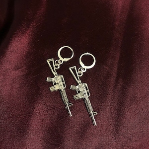 May include: A pair of silver-colored earrings featuring a rifle design. The earrings have a lever back closure and are displayed against a dark red background. The rifle design is detailed and has a metallic finish.