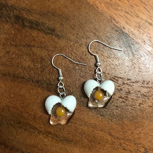 May include: A pair of silver dangle earrings with a white and yellow egg yolk design. The egg is cracked open and the yolk is visible.