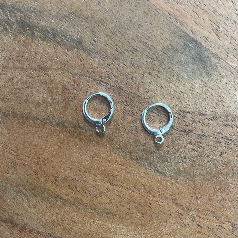 Peut inclure: Deux fermoirs de boucles d'oreilles argent&eacute;s, chacun avec un design circulaire et une petite boucle en bas. Les fermoirs sont pos&eacute;s sur une surface en bois, mettant en valeur leur design simple mais &eacute;l&eacute;gant.