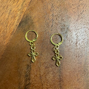 Könnte beinhalten: Ein Paar goldene Creolen mit einem kleinen goldenen Kreuzanhänger an jedem Ohrring.