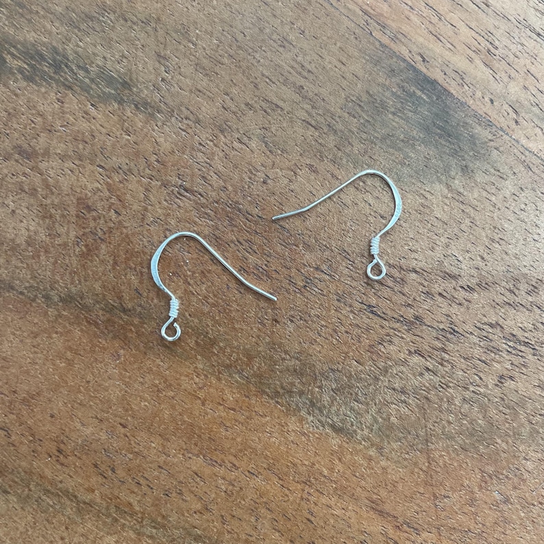 Peut inclure: Deux crochets de boucles d'oreilles argent&eacute;s, pr&eacute;sent&eacute;s sur une surface en bois. Les crochets ont un design simple et incurv&eacute; avec une petite boucle &agrave; la base pour attacher les composants des boucles d'oreilles. Les crochets sont en m&eacute;tal brillant.
