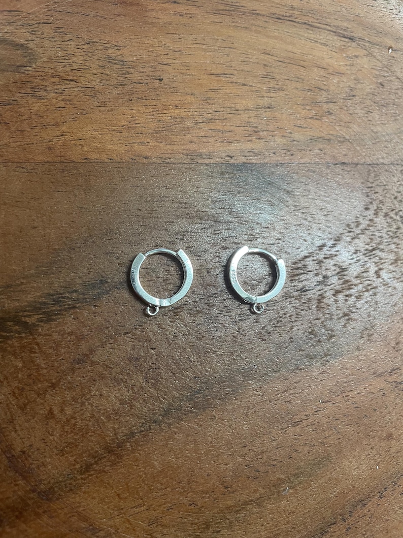 Peut inclure: Deux petites boucles d'oreilles argent&eacute;es en forme de cr&eacute;oles. Chaque boucle d'oreille pr&eacute;sente un design circulaire simple avec une petite boucle en bas. Les boucles d'oreilles sont pos&eacute;es sur une surface en bois.