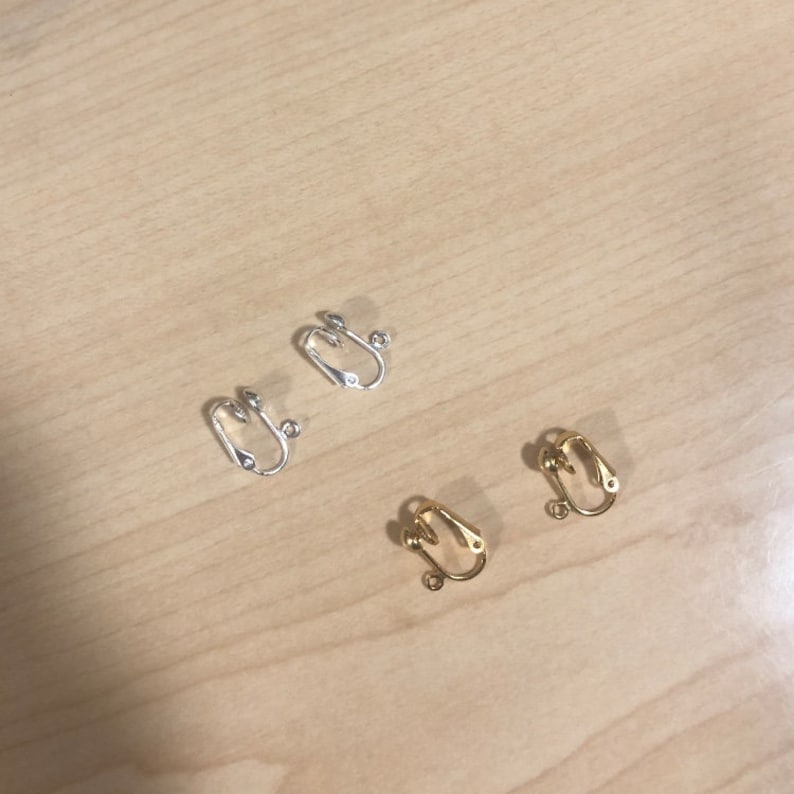 Peut inclure: Quatre attaches de boucles d'oreilles &agrave; clip, argent&eacute;es et dor&eacute;es, dispos&eacute;es sur une surface en bois clair. Les attaches ont un design simple et incurv&eacute; avec une petite boucle pour fixer les boucles d'oreilles.